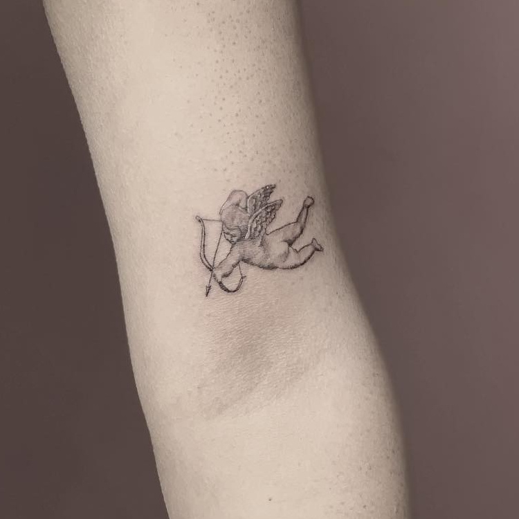 Fine-line cherub tattoo drawing a bow and arrow, on the back of the upper arm