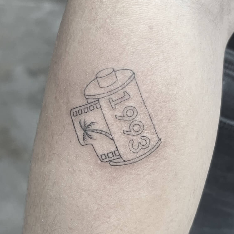 Fine-line 35mm film canister tattoo dated 1993, with palm trees on the film strip, on the forearm