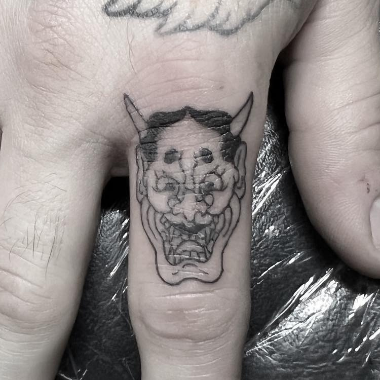 Black-outline oni demon mask tattoo with horns, down the middle finger