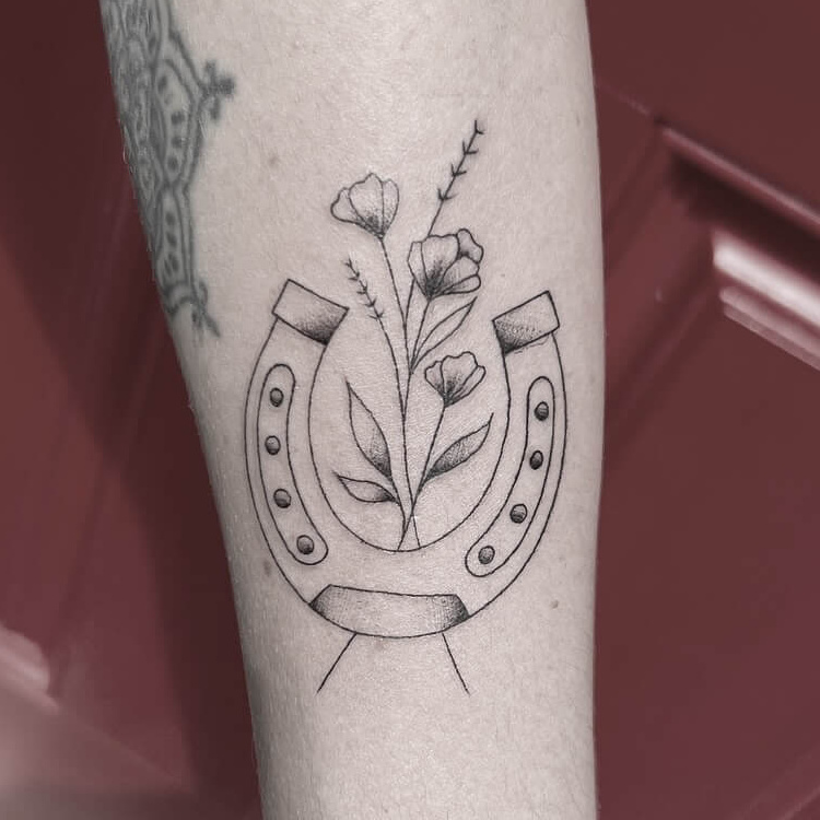 Fine-line horseshoe tattoo with wildflowers growing through it, on the forearm